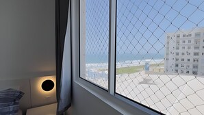 A room with a window overlooking a chain-link fence and a building across the street.