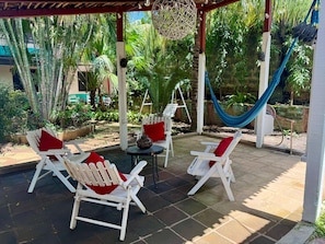 A covered outdoor seating area with white chairs and red cushions, surrounded by lush greenery and a hammock. - Hostal Harvest House (León)