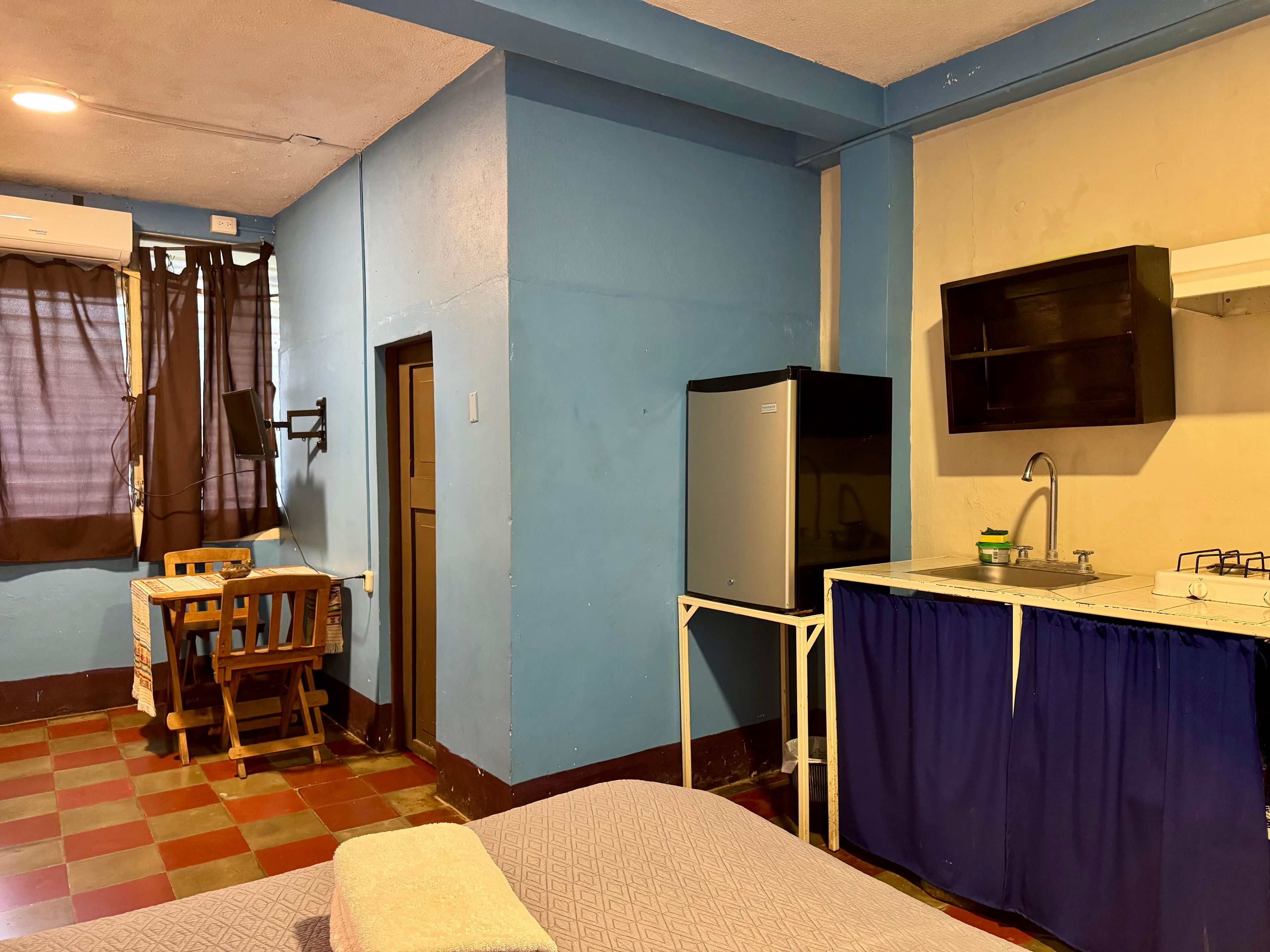 A compact room with a kitchenette, a single bed, a chair, and a window with curtains.