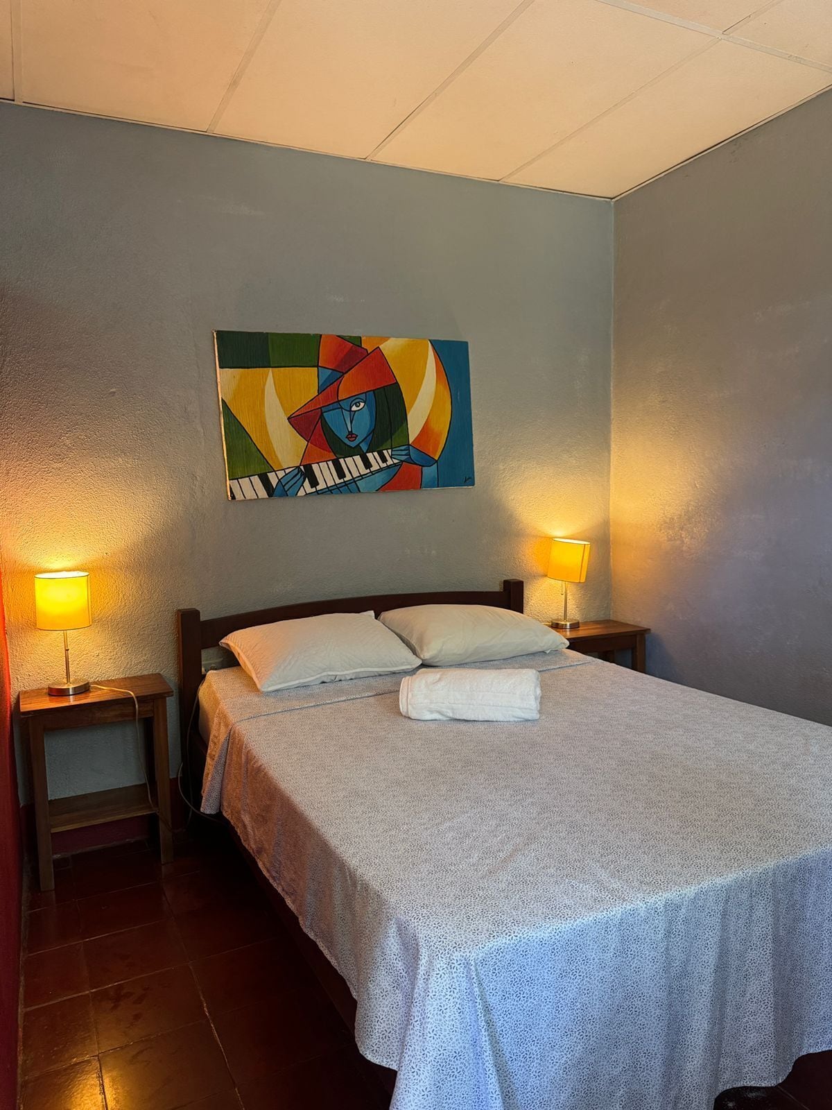 Quarto de hotel com cama, duas luminárias de cabeceira, um quadro na parede e uma mesinha de cabeceira de madeira.