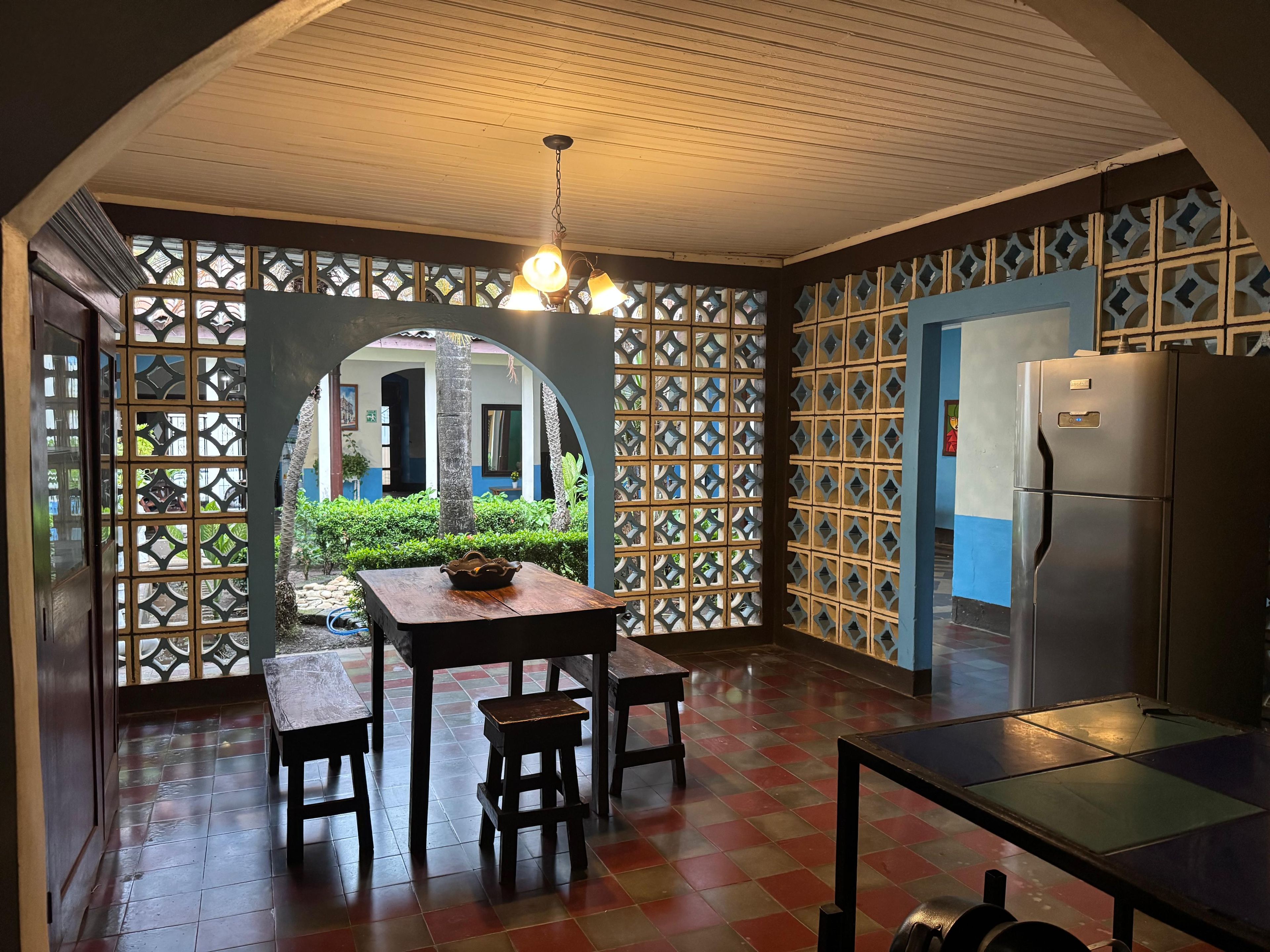 Una cocina con una mesa de madera, sillas, un refrigerador y vistas a un espacio exterior a través de un separador decorativo.