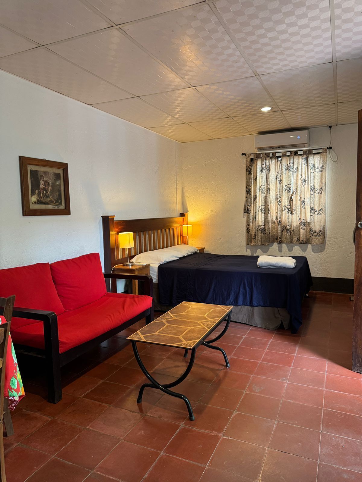 A hotel room with a red sofa, a bed with a dark blue bedspread, a small table, and an air conditioning unit.