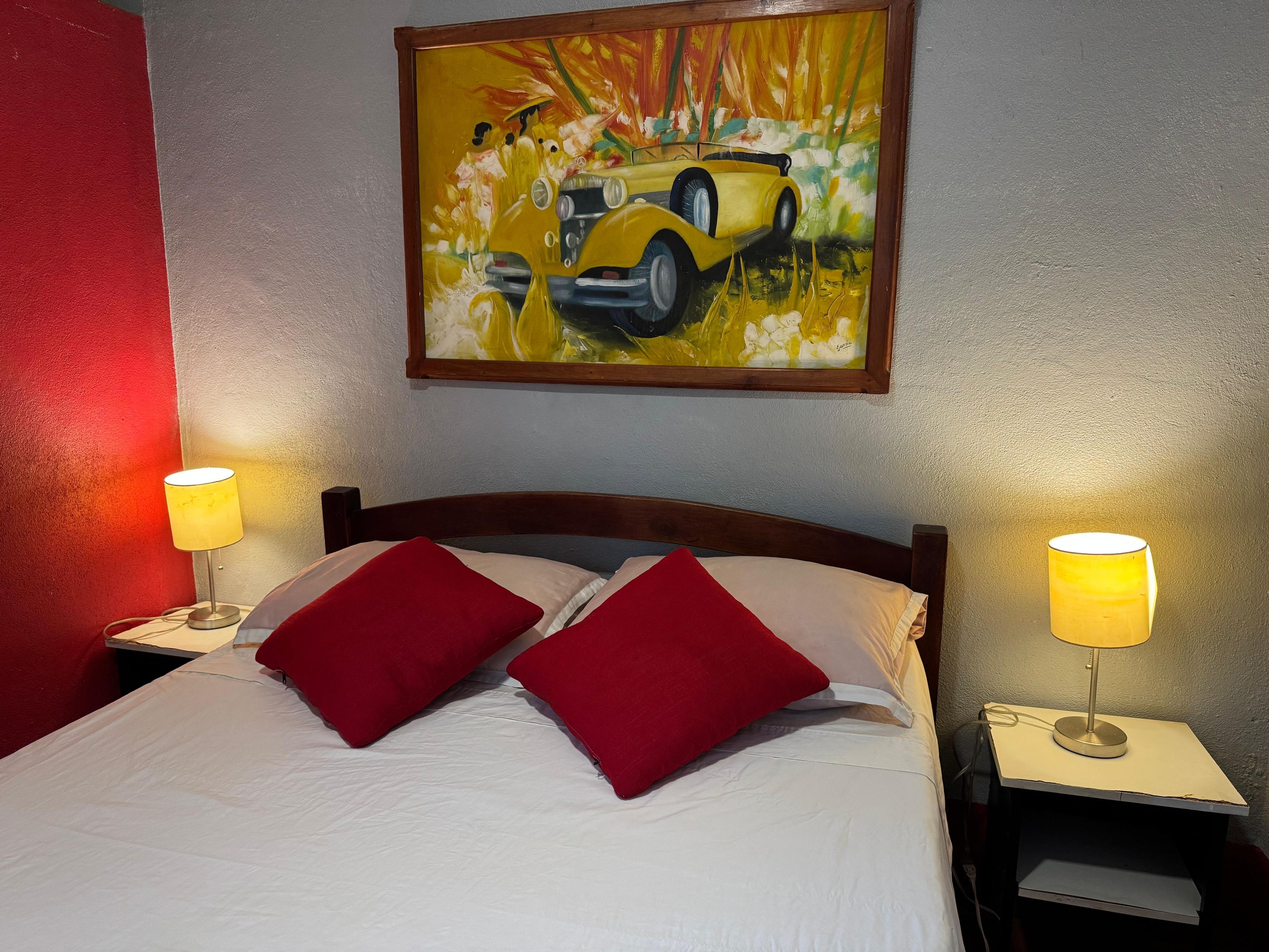 A hotel room with a bed, two red pillows, a painting of a yellow car, and two bedside lamps.