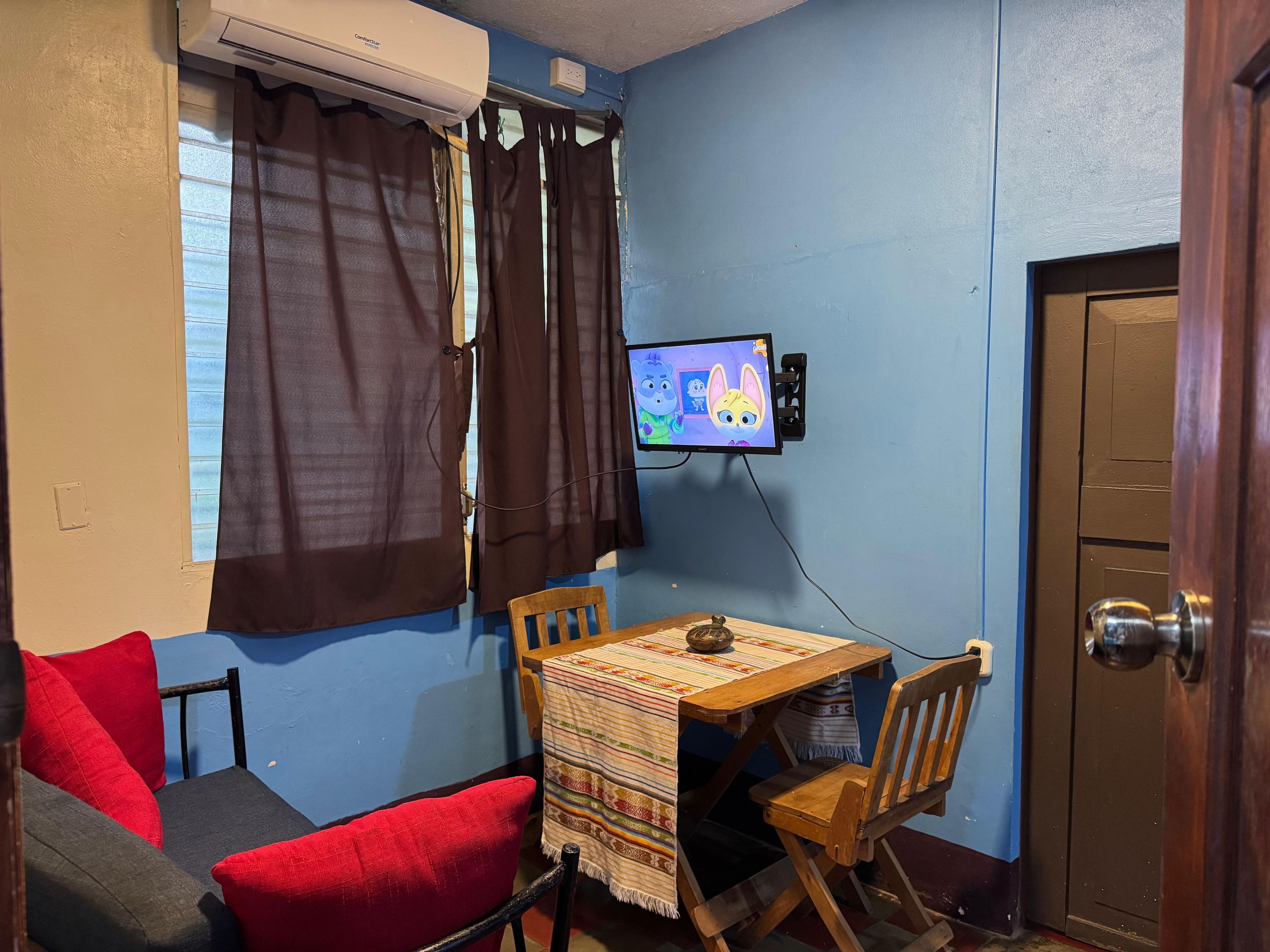 A small room with a blue wall, a window with curtains, a television mounted on the wall, a wooden table with a colorful tablecloth, and a door.