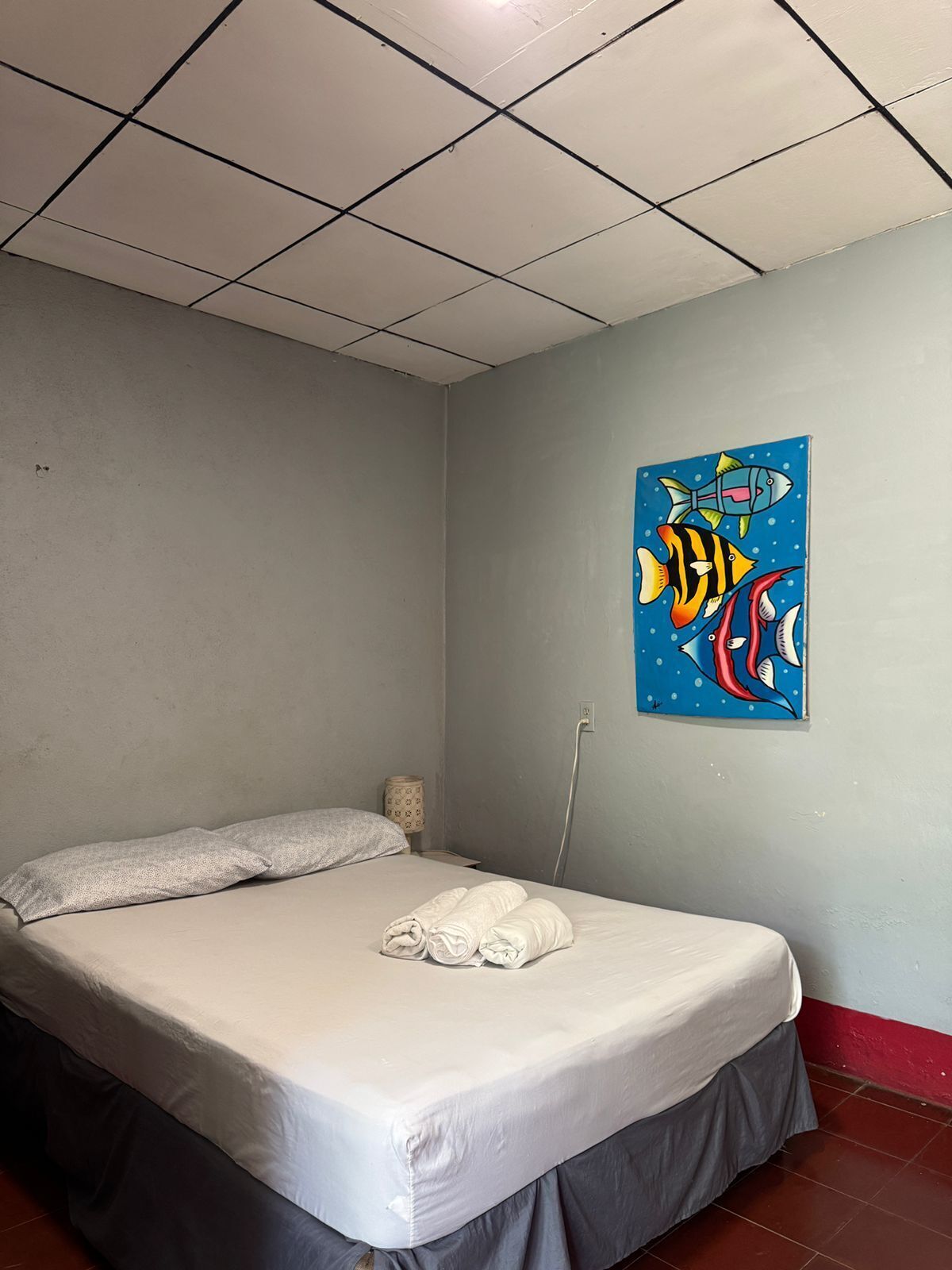 A single bed with a grey skirt, a grey pillow, and a blue wall with a fish painting.
