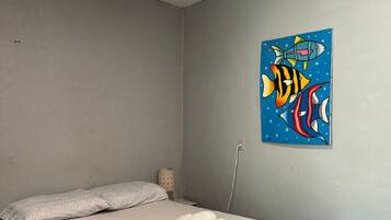 A single bed with a grey skirt, a grey pillow, and a blue wall with a fish painting.