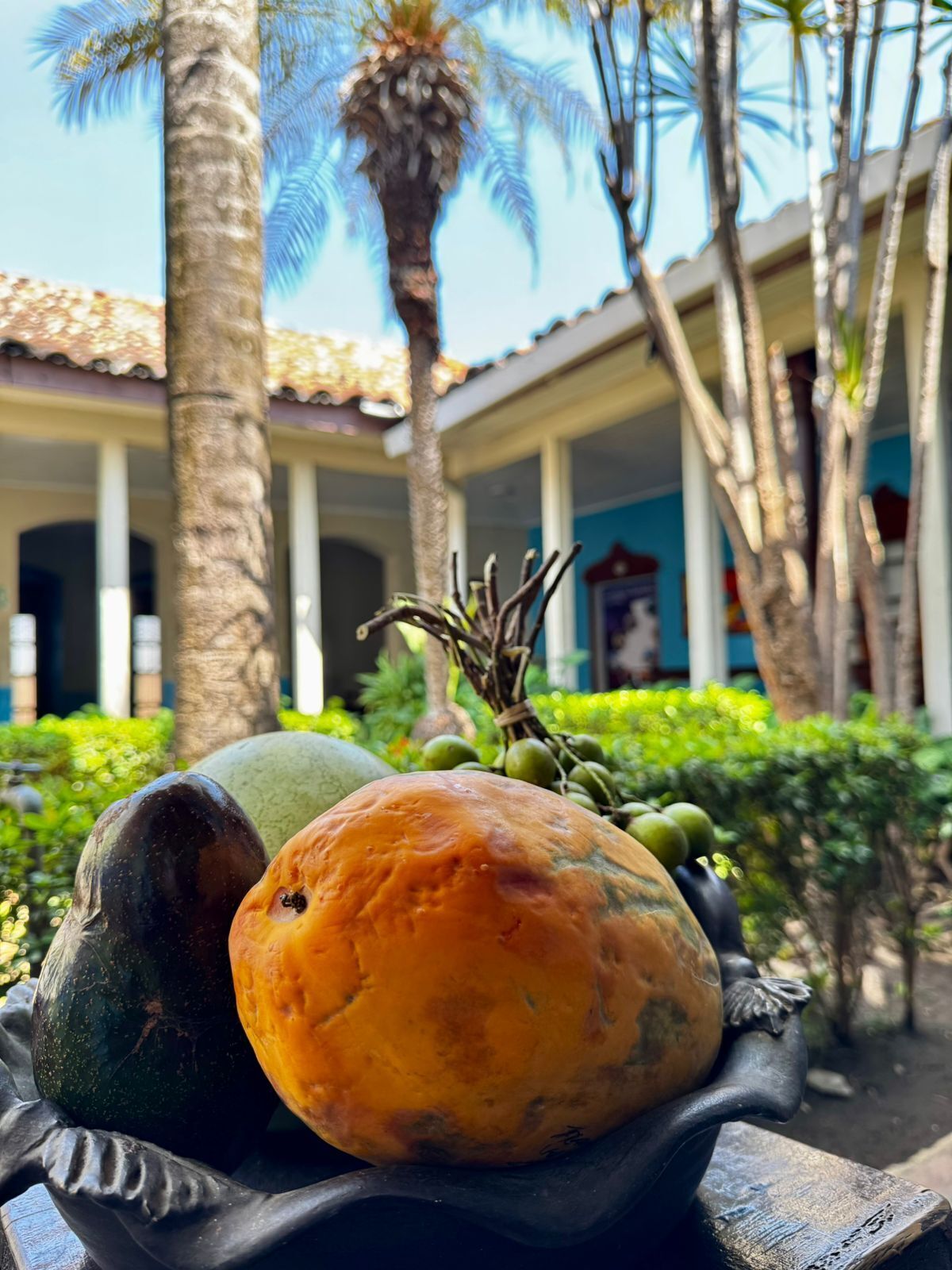 Un frutero con diversas frutas sobre una mesa, palmeras y un edificio con columnas al fondo.