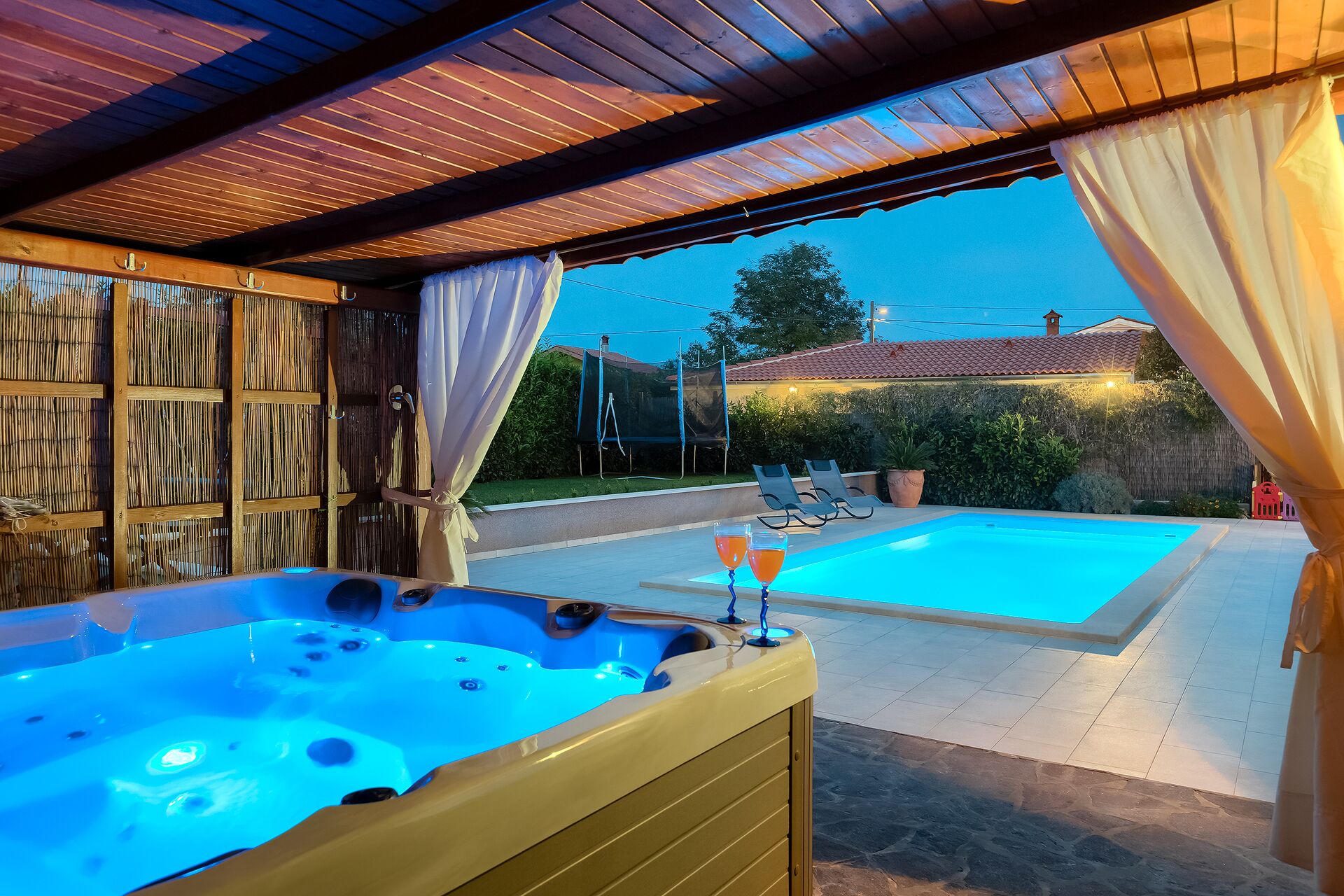 A hot tub and a swimming pool under a wooden pergola with curtains.
