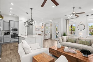 A modern kitchen with white cabinets, a central island, and a dining area with a white sofa and wooden coffee table. - Pelican Paradise a coastal townhome on 30A~premier location, steps to pool, LSV (Watersound)