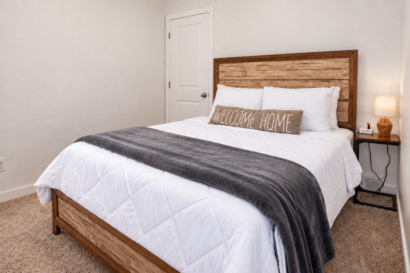 A bedroom with a wooden headboard, a white bedspread, a gray throw blanket, and a 'WELCOME HOME' pillow.