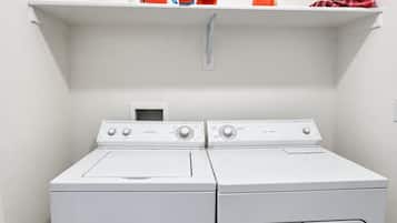 A laundry area with two white front-loading washing machines and a white shelf above them holding various items.