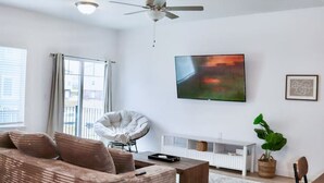 Ein modernes Wohnzimmer mit Sofa, Sessel, Esstisch, Deckenventilator, Flachbildfernseher und einer Topfpflanze.