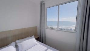 A hotel room with a bed, a wooden headboard, a window with a view of the sea, and a sign on the window. - Solarium Beach Club -Apartamento T1.1003 (Itapoá)