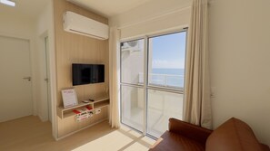 Een moderne hotelkamer met een televisie, een klein plankje met spullen, een glazen schuifdeur naar een balkon en een airconditioning.