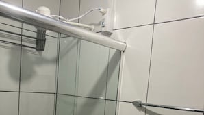 En brusekabine med glasvægge, et metalgreb, hvide fliser og en synlig stikkontakt.