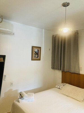 Ein Hotelzimmer mit Bett, einem kleinen Tisch mit einem Handtuch, einem gerahmten Bild an der Wand, einem Fenster mit Vorhängen und einer Klimaanlage.