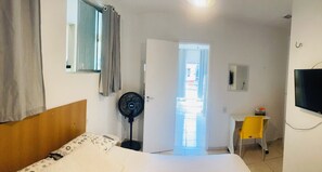 A hotel room with a bed, a desk with a chair, a TV, a fan, and a door leading to an outdoor area. - Complete apartment behind the Shopping Mall (Belém)
