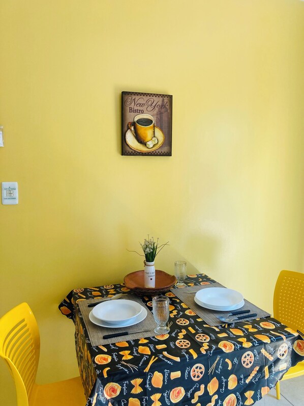 Ein Esstisch mit schwarzem Tischläufer, der Pasta-Muster zeigt, zwei weiße Teller, eine Vase mit Blumen und ein Gemälde einer Kaffeetasse an der Wand.