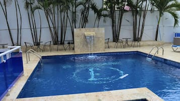 Una piscina rettangolare con bordo piastrellato, una piccola cascata e palme circostanti.