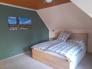 Ein Schlafzimmer mit schrägen Wänden, einem Bett mit kariertem Überwurf, einem Nachttisch und einer Wandlampe.
