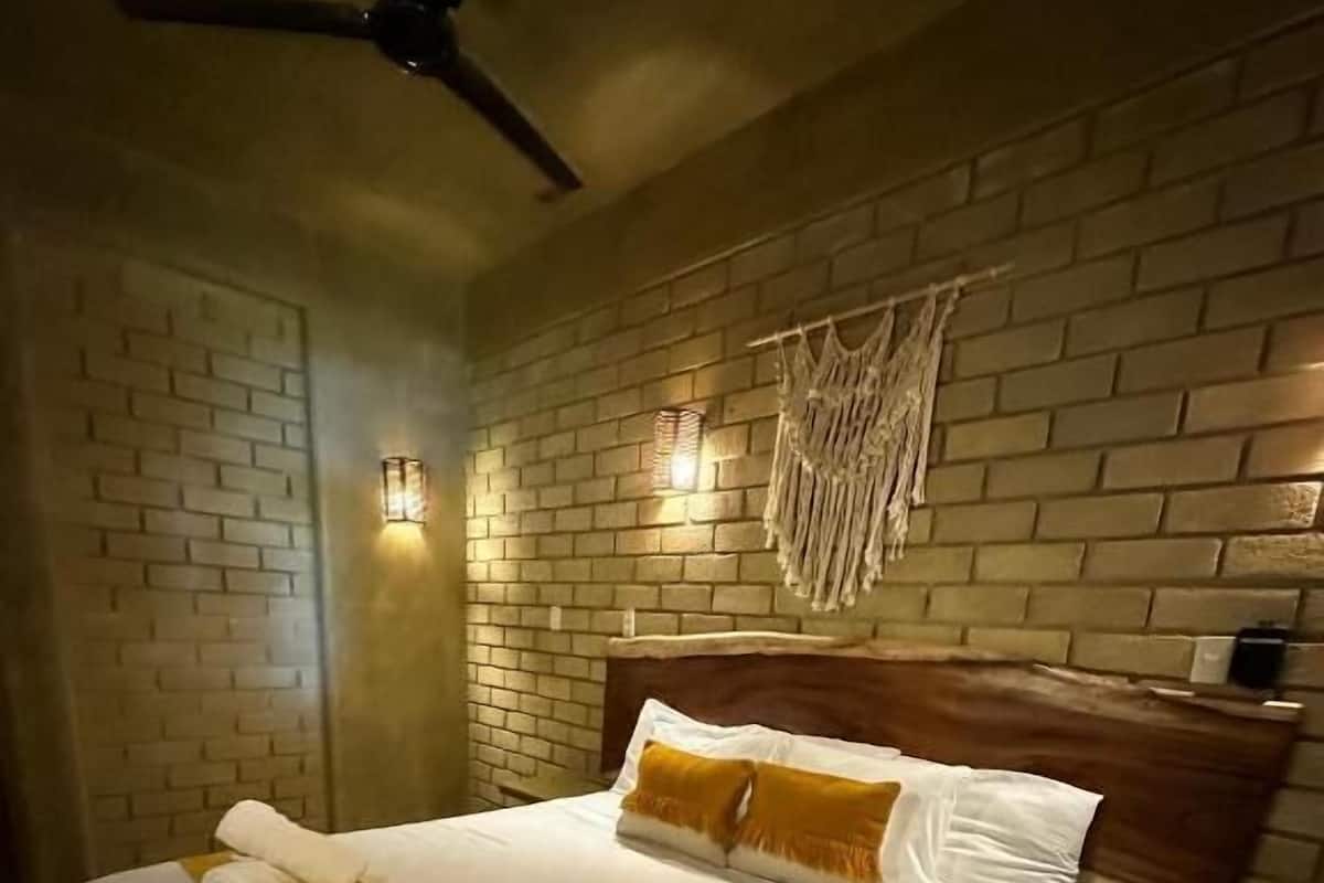 Une chambre d’hôtel avec un mur en briques, un lit avec une literie blanche et un élément décoratif jaune, un ventilateur de plafond et une décoration murale en macramé.