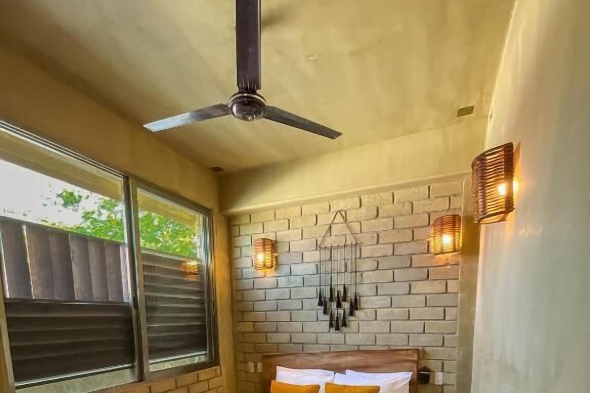 Quarto com parede de tijolos, cama com roupa de cama branca e detalhes em laranja, ventilador de teto e luminárias fixadas na parede.