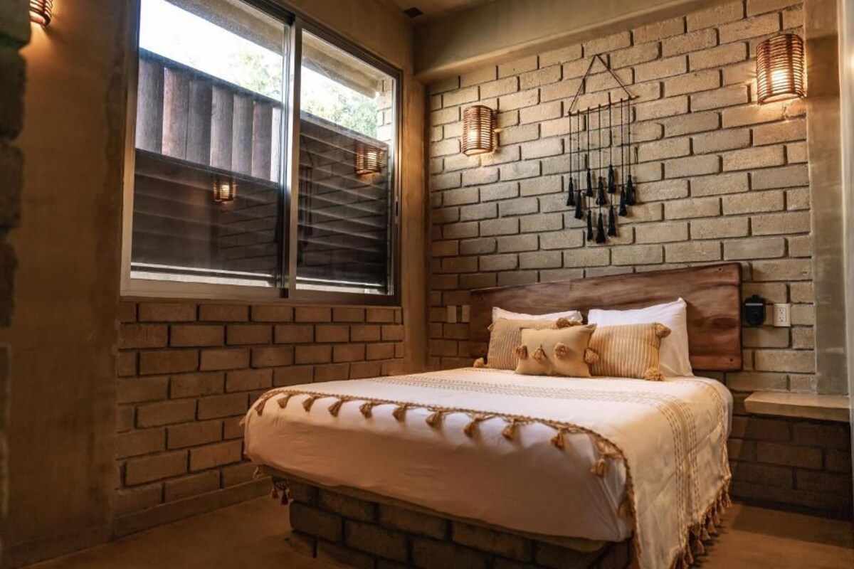 Quarto com parede de tijolos, cabeceira de madeira, cama com roupa de cama branca e duas luminárias fixadas na parede.