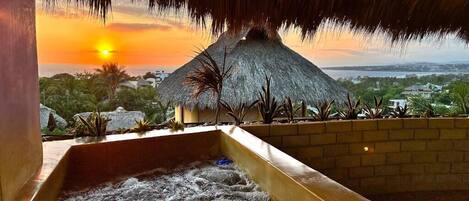 Un jacuzzi con vista a un atardecer y una estructura de techo de paja.