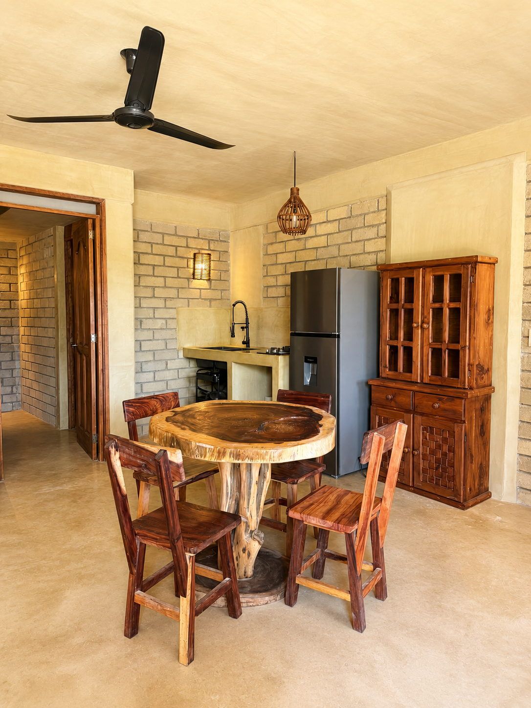 A dining area with a round wooden table surrounded by chairs, a ceiling fan, a refrigerator, and a wooden cabinet.