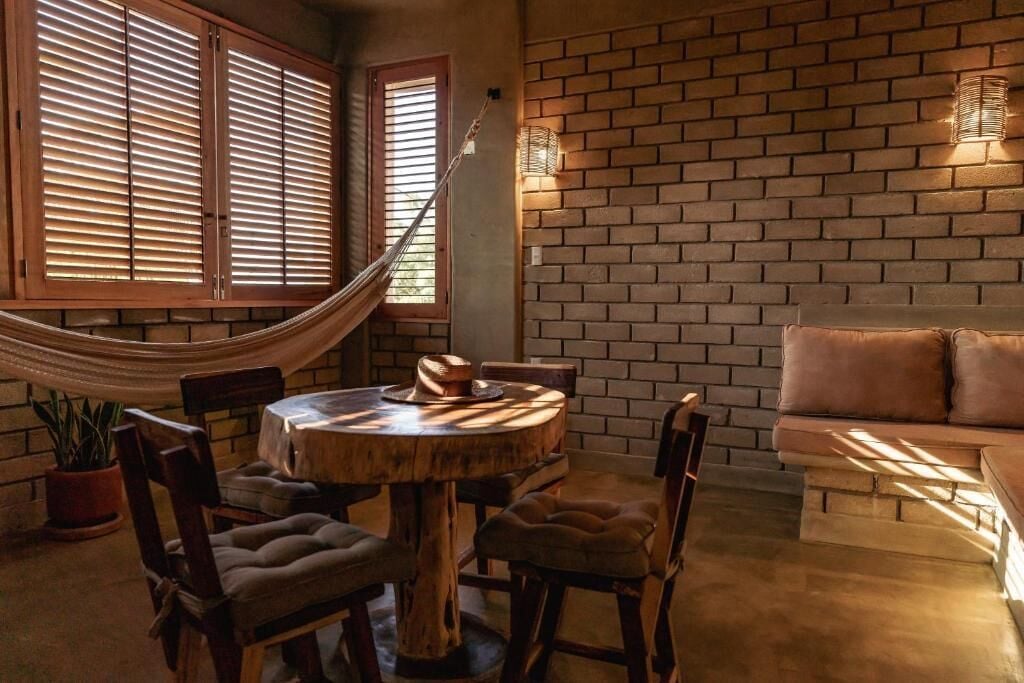 Un cuarto acogedor con una mesa redonda de madera, sillas con acolchado, una hamaca y una pared de ladrillo con una lámpara.
