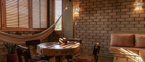 Un cuarto acogedor con una mesa redonda de madera, sillas con acolchado, una hamaca y una pared de ladrillo con una lámpara.