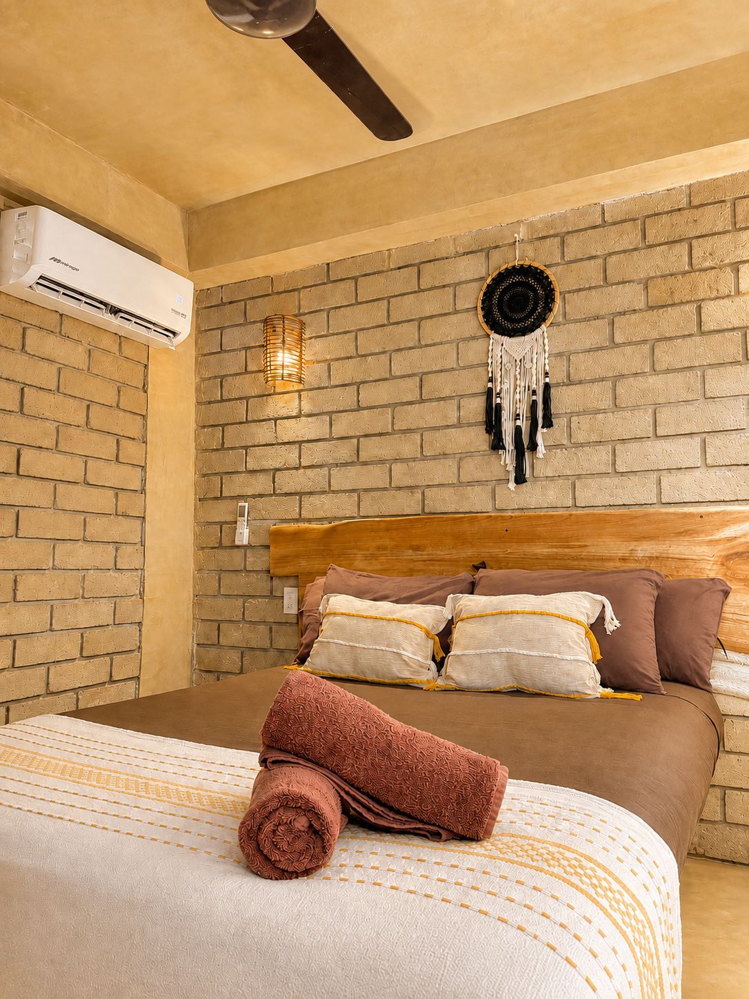 A bedroom with a brick wall, a wooden headboard, and a bed with multiple pillows and a rolled-up towel.