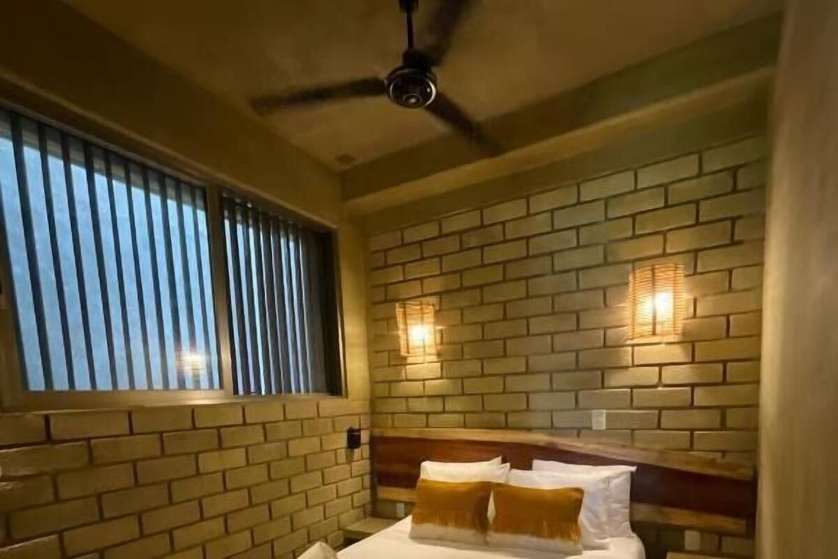 Une chambre d’hôtel avec un mur en briques, un lit avec une literie blanche, une tête de lit en bois et un ventilateur de plafond.