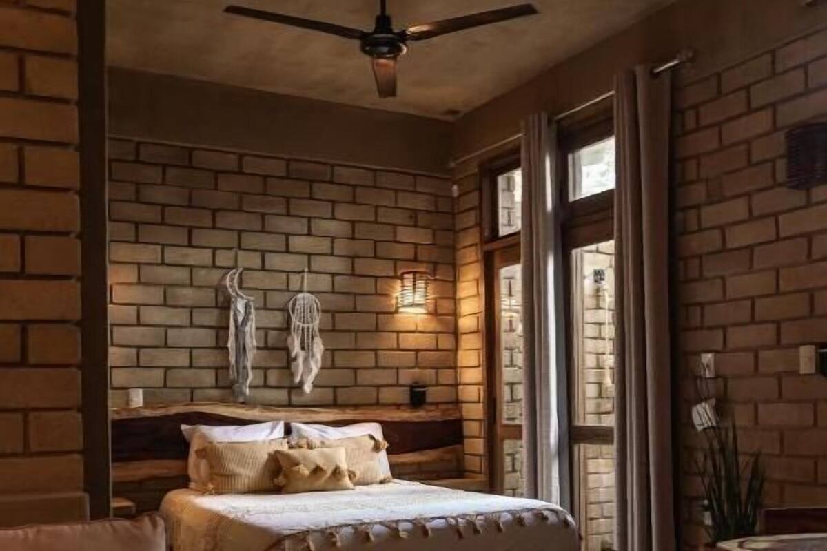 Une chambre avec un mur en briques, un lit avec une tête de lit, un ventilateur de plafond et une fenêtre avec des rideaux.