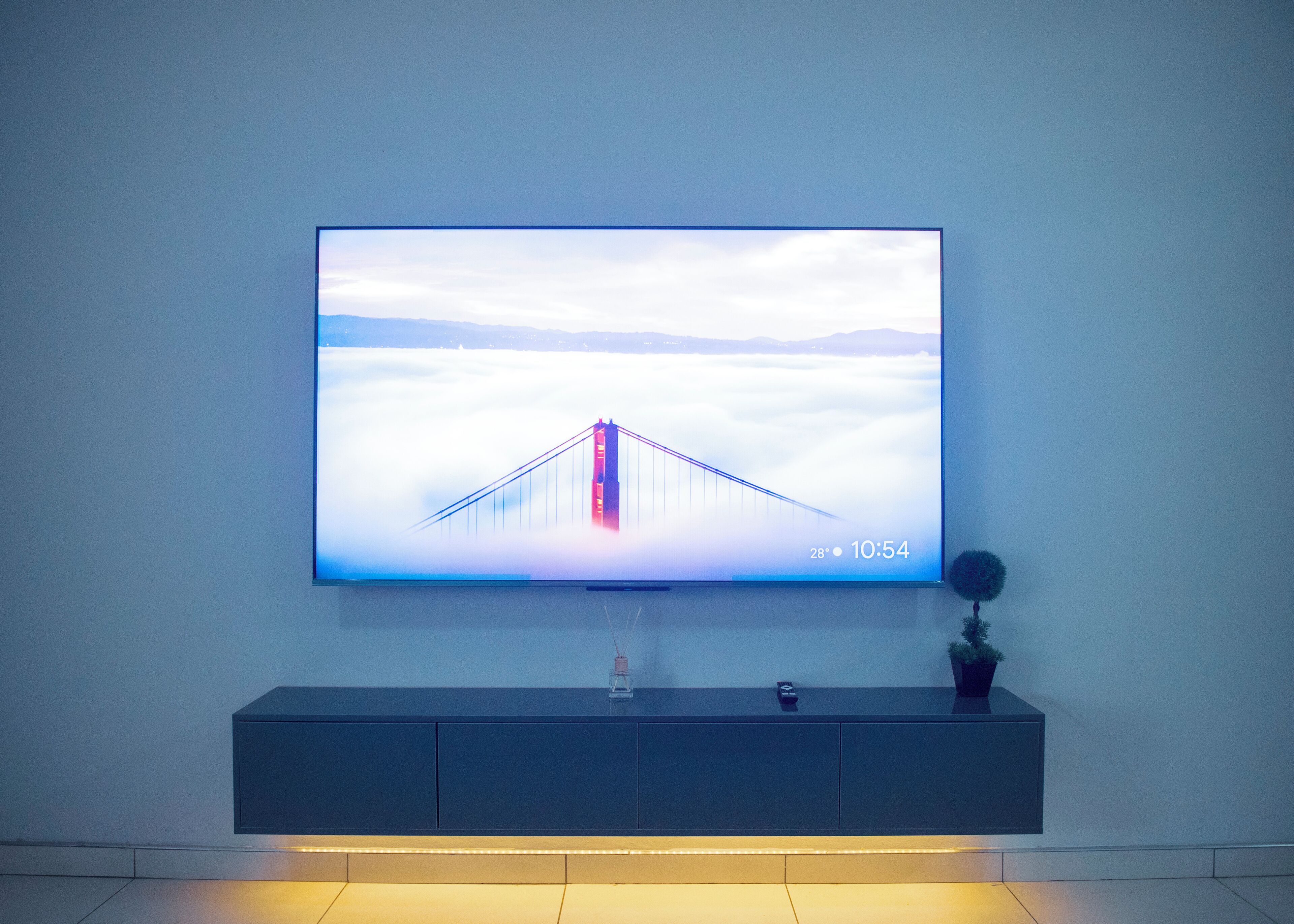 Ein Flachbildfernseher an der Wand, der eine Brücke über verschwommenes Gelände zeigt, mit einer digitalen Uhr, die 10:54 Uhr anzeigt und eine Temperatur von 28 Grad anzeigt.