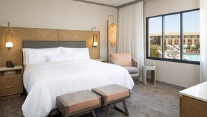Ein Hotelzimmer mit einem großen Bett, Nachttischlampen, einem Sessel, einem kleinen Tisch und Blick auf ein Gebäude und Wasser.