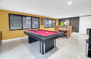 A pool table in a room with yellow walls, a dining area with wooden furniture, and large windows with blinds. - El bosque encantado (Miami)