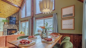 A dining area with a wooden table set for four, surrounded by green upholstered chairs. A stone fireplace with a fire is visible in the background, and there are framed pictures on the walls.