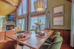 A dining area with a wooden table set for four, surrounded by green upholstered chairs. A stone fireplace with a fire is visible in the background, and there are framed pictures on the walls. - Smoky Mountain Views - Fire Pit, Sauna & Hot Tub (Bryson City)