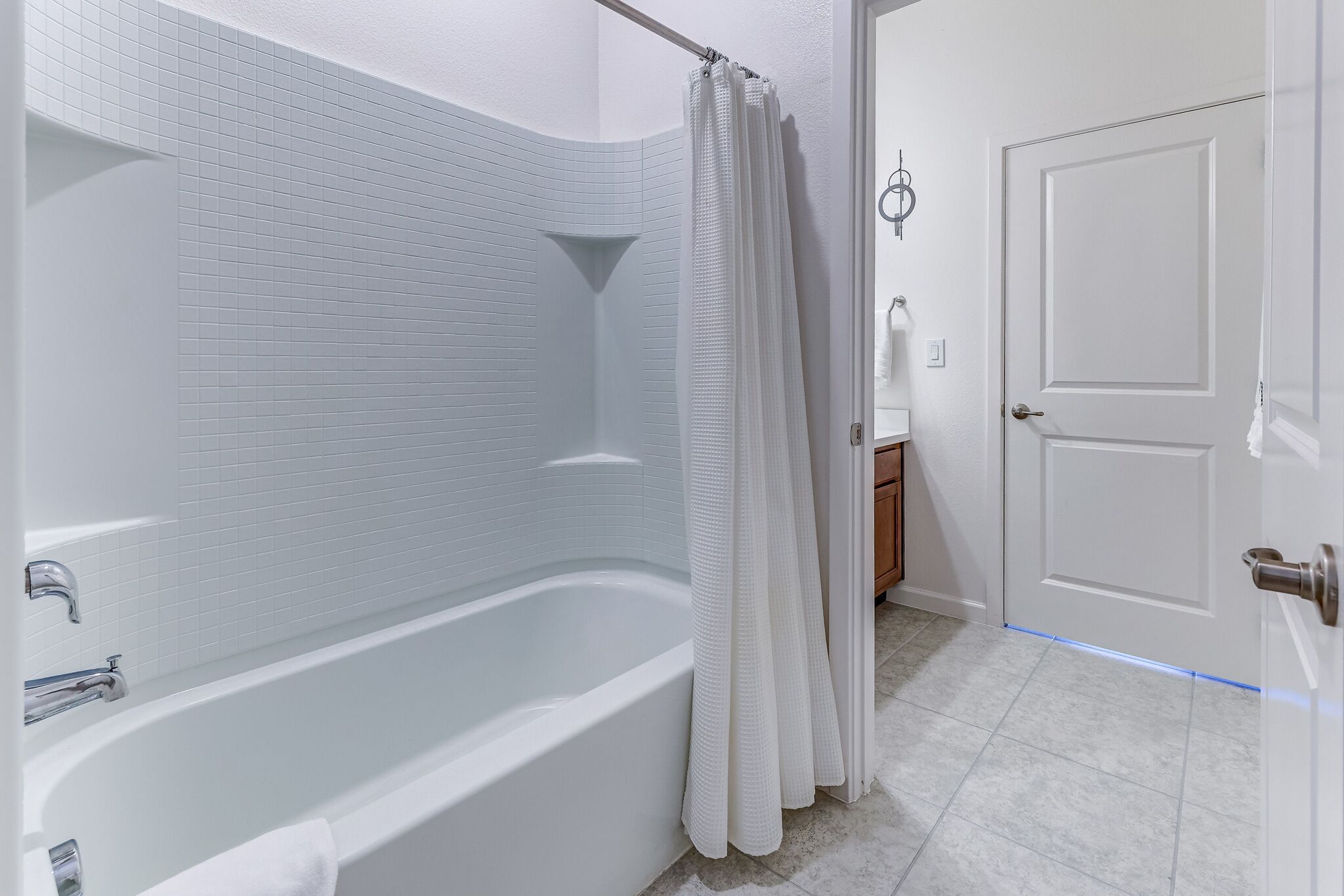 Une salle de bain moderne avec une baignoire, des murs recouverts de carreaux blancs, un rideau de douche et une porte dotée d’un interrupteur.