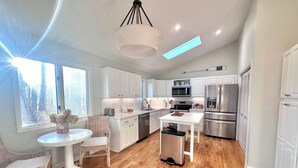 Une cuisine moderne avec des meubles blancs, des appareils en acier inoxydable, un coin repas avec une table ronde et des chaises, et une vue sur l’extérieur grâce à de grandes fenêtres.