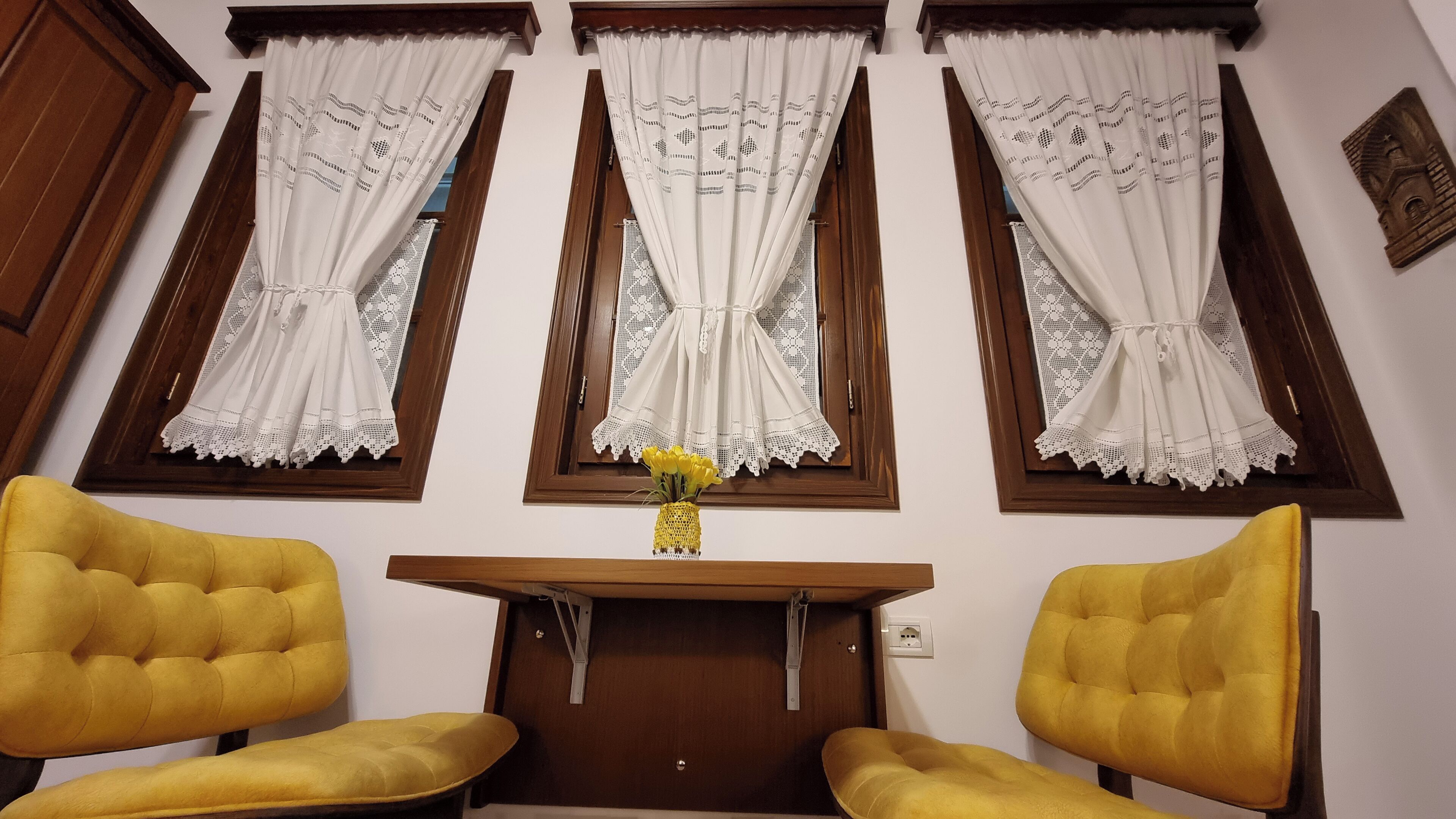 Une pièce dotée de trois fenêtres, chacune ornée de rideaux blancs à motifs de dentelle. On y trouve deux fauteuils jaunes et une petite table en bois sur laquelle est posé un vase rempli de fleurs jaunes.