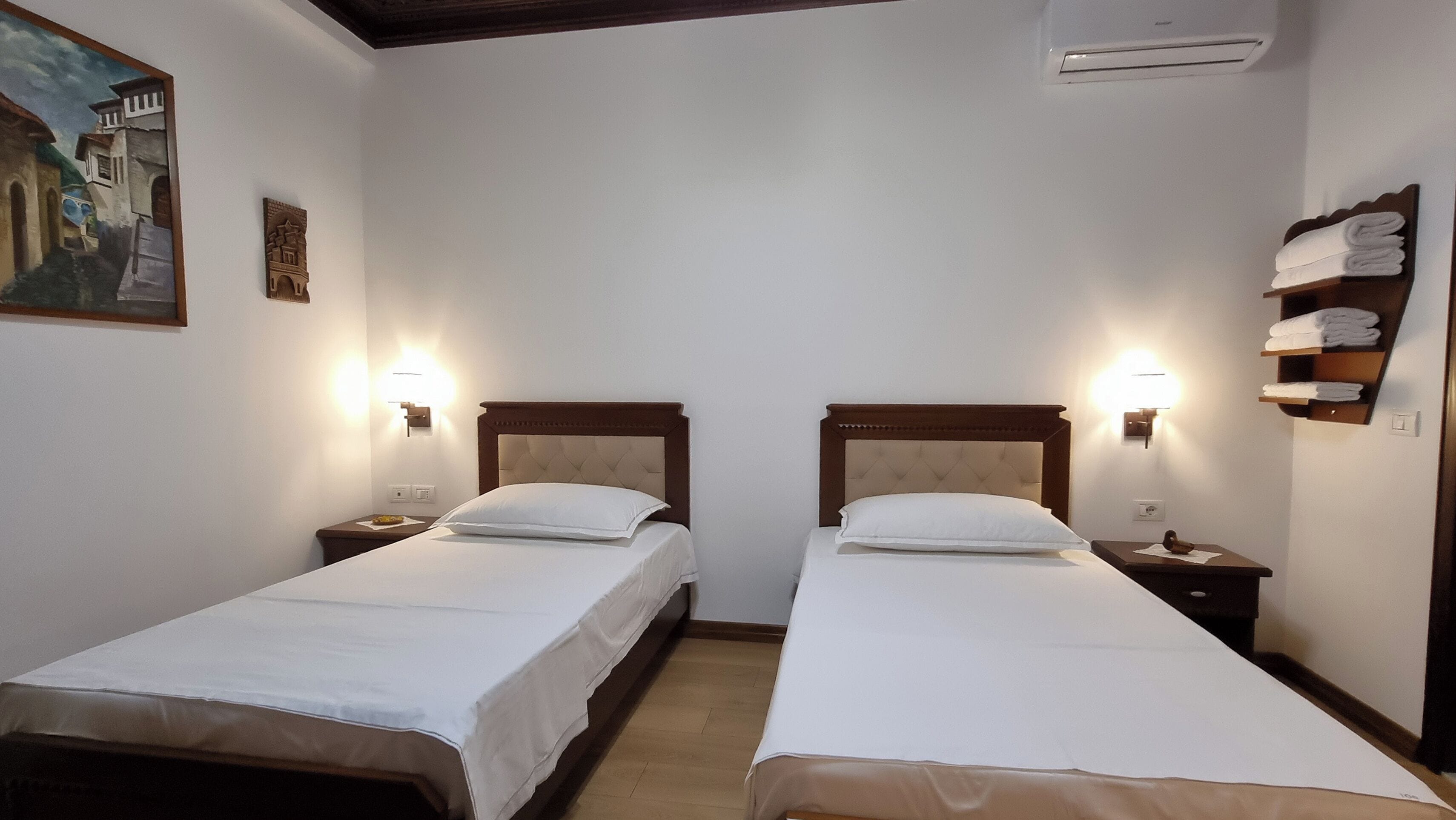 Ett hotellrum med två separata sängar, var och en med ett träfärgat sänggavel och vitt sänglinne. På vardera sidan av sängarna finns väggmonterade lampor, och till höger om sängarna står en hylla med hopvikta handdukar.
