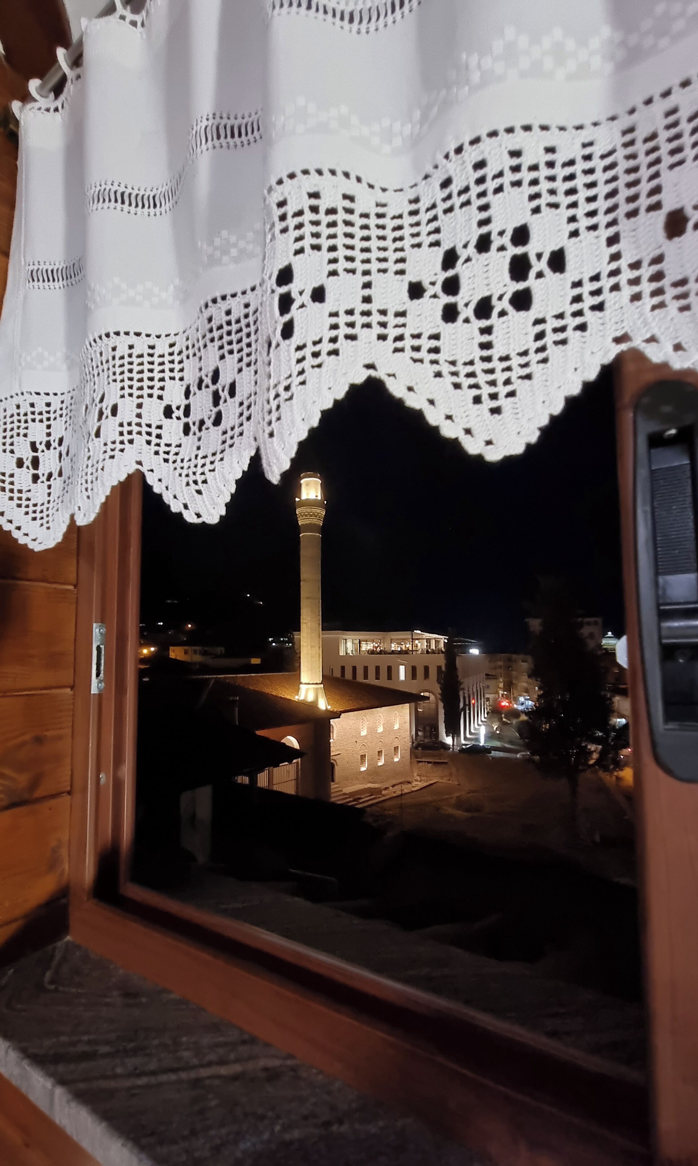 A view through a window with a white lace curtain, a wooden door, and a glimpse of a building with a lit tower at night.