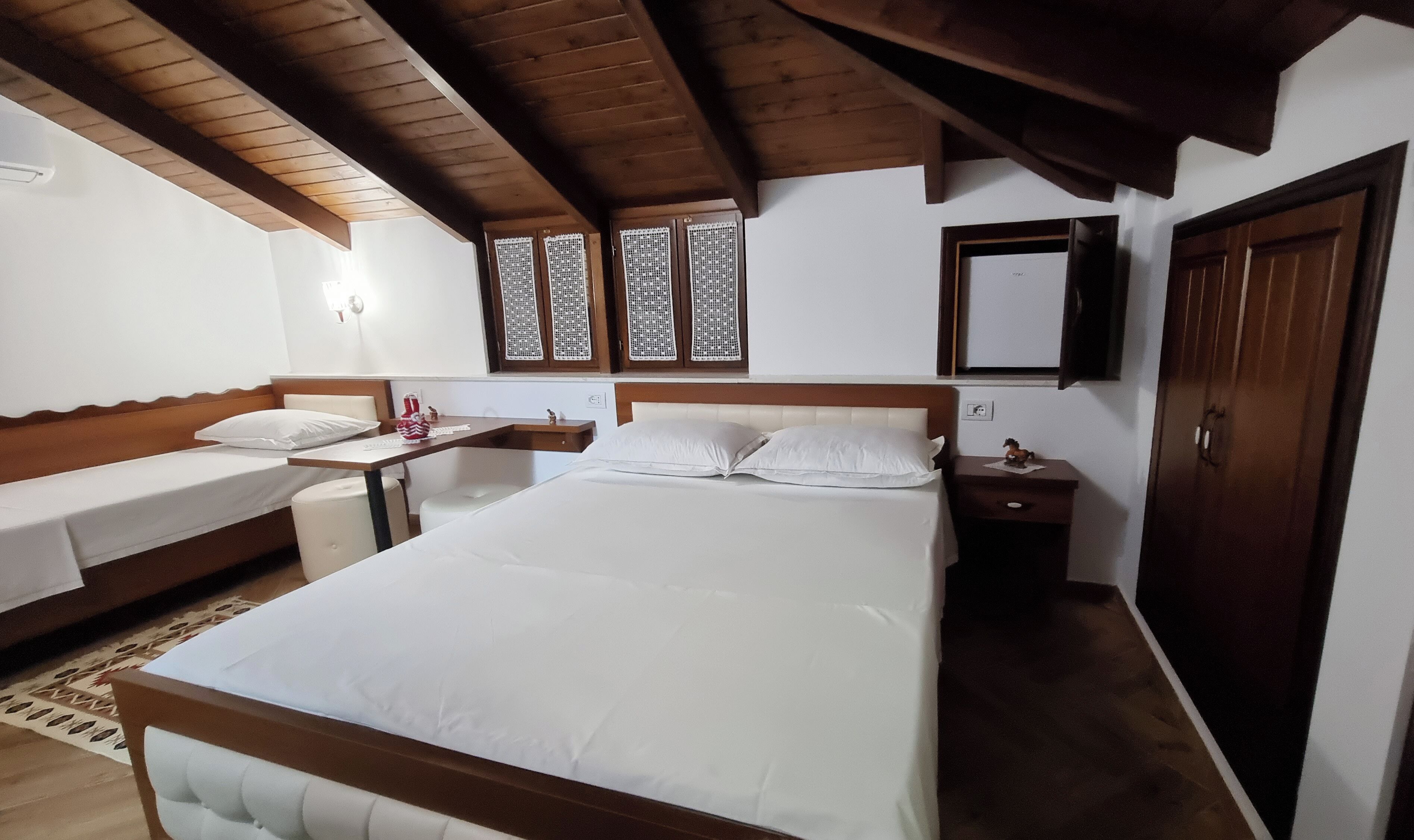 A hotel room with a large bed, a smaller bed, a desk, a chair, a wardrobe, and a window with decorative shutters.