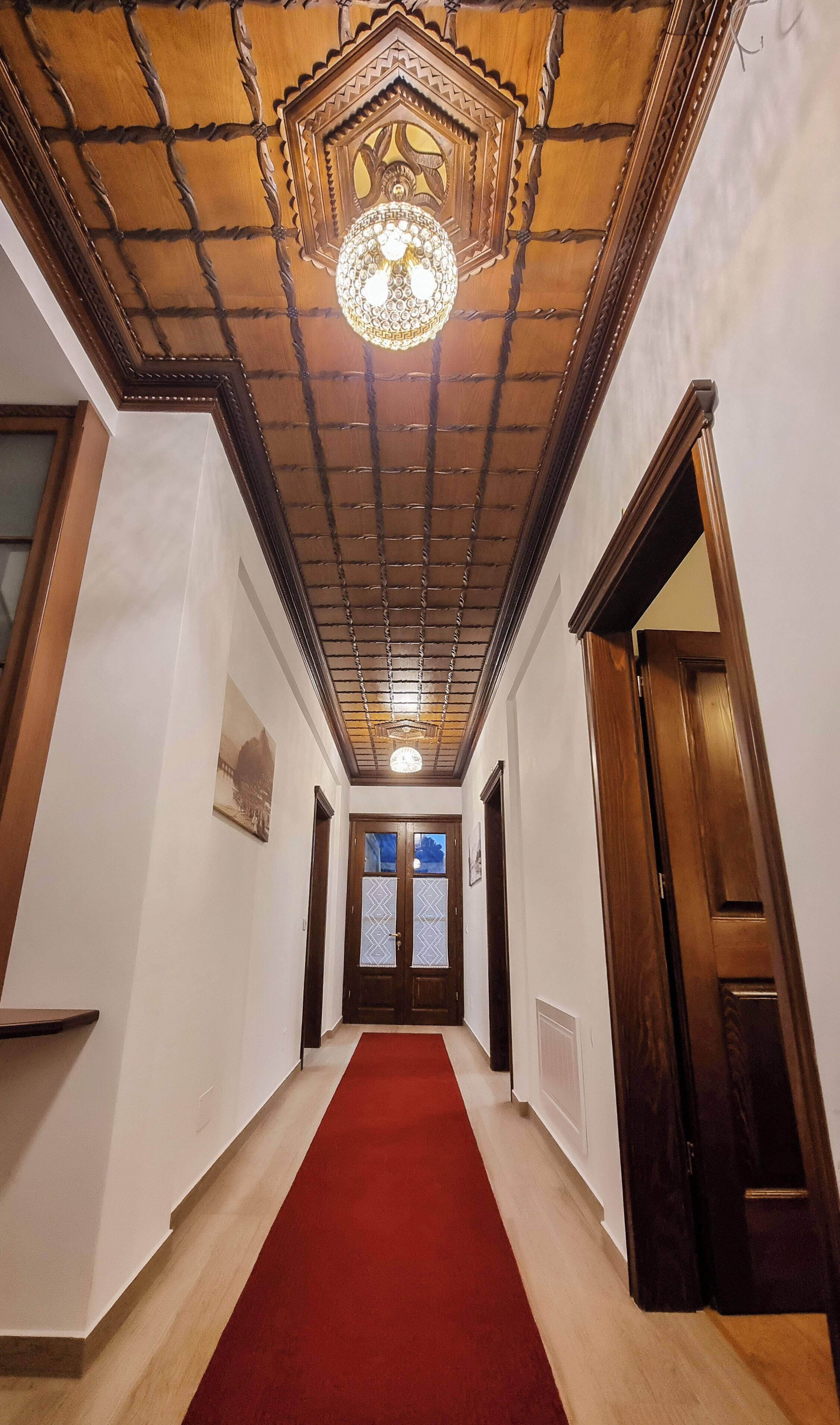 A hallway with a red carpet, wooden doors, and a decorative ceiling with a chandelier.