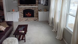 A living room with a fireplace, a flat-screen TV mounted above it, a sofa, a coffee table, and a window with curtains.