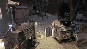 A deck with a grill and a barbecue, surrounded by a snow-covered yard and trees.