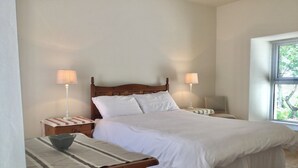 A neatly made bed with white linens, two bedside lamps, a wooden nightstand, and a window with blinds. - Delightful charming townhouse in seaside Ballyvaughan awaits you (Ballyvaghan)