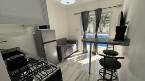 Eine kleine Küche mit Gasherd, Mikrowelle und Kühlschrank. Ein graues Sofa und eine kleine Bar mit einem Stuhl sind zu sehen. Ein großes Fenster bietet Blick auf einen Swimmingpool.